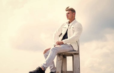 Hem gündelik hem de modaya uygun giyinmek. Yakışıklı adam açık havada rahat giyiniyor. Adam bulutlu gökyüzünde sandalyeye oturur. Günlük kıyafet dolabı. Moda trendleri. Moda tarzı. Erkek giyim mağazası. Günlük giysiler.