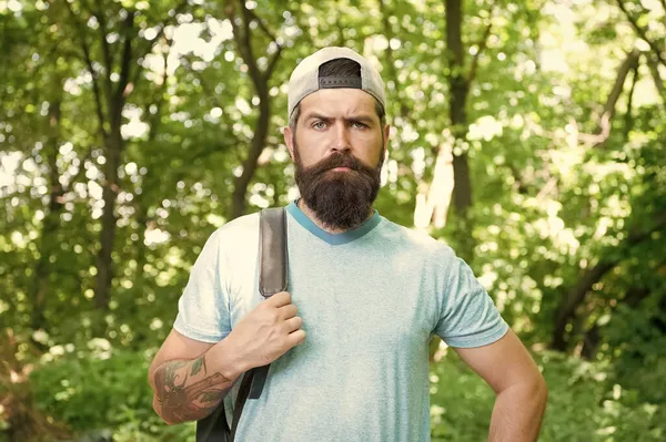 Зрілий бородатий чоловік з бородою і вусами в літній сорочці і кепці тримає рюкзак в лісі, мандрівник — стокове фото