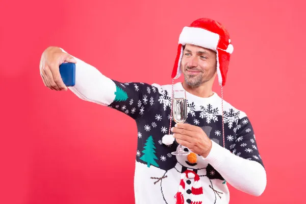Homem positivo em camisola de inverno e chapéu fazendo selfie no smartphone enquanto bebe champanhe, xmas. — Fotografia de Stock