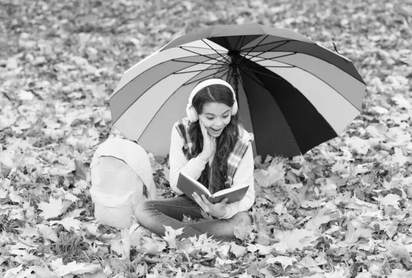 Şaşkın çocuk kulaklıkla müzik dinlerken sonbahar ormanında renkli şemsiyeli yaprakların arasında oturmuş okul çantası, kitap, eğitim okuyordu. — Stok fotoğraf