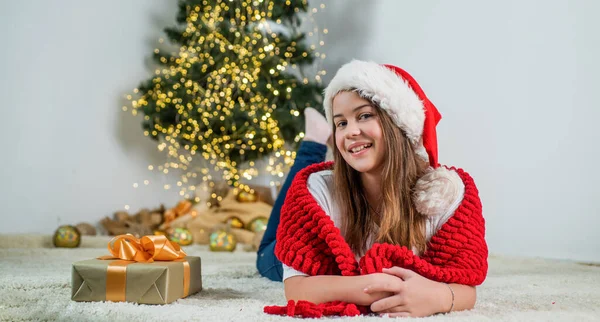 메리 크리스마스. 쾌활 한 소녀는 산타 모자를 쓰고 있다. 스카프를 두르고 있는 꼬마. 집에서 크리스마스 장식을 하고 있는아이. 어린 시절의 행복. 겨울 방학 과 휴가. 준비하고 축하 해. 새해 복 많이 받으세요 — 스톡 사진