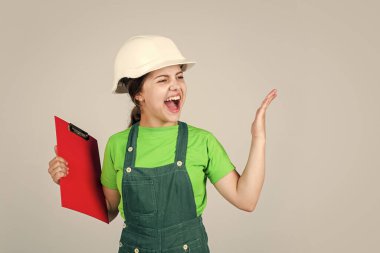 Sert şapkalı kızgın çocuk not alıyor. Çocuk tutulan belge klasörü. Çocuk, güvenlik şapkalı inşaat işçisi olmayı öğreniyor. Kasklı, tabletli küçük çocuk. Onarım planlaması. kızgınlığını ifade et