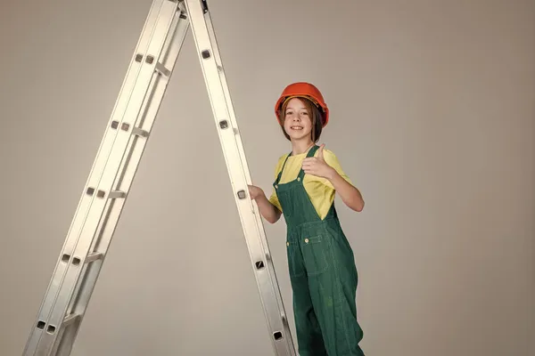 Allegra ragazza adolescente in uniforme da lavoro e casco sulla scala spettacolo pollice in su, edificio — Foto Stock