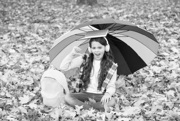 Parfait jour d'automne de l'enfant gai sous parapluie coloré avec sac d'école écouter de la musique dans les écouteurs et tenir le livre dans le parc de la saison d'automne profiter du beau temps, journée de la connaissance — Photo