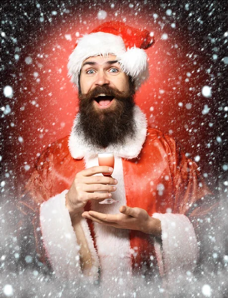 Schöner bärtiger Weihnachtsmann mit langem Bart auf lächelndem Gesicht mit einem Glas alkoholischen Schnaps in der Hand in Weihnachts- oder Weihnachtspullover und Neujahrsmütze auf rotem Studiohintergrund — Stockfoto