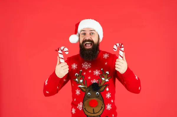 Sorprendido barbudo chico en santa claus sombrero y suéter celebrar caramelo palo sobre fondo rojo, año nuevo —  Fotos de Stock