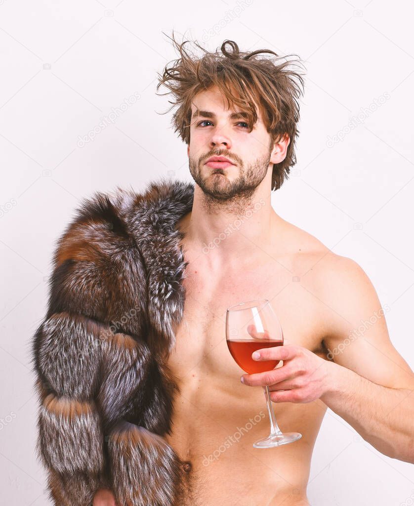 Sexy sleepy macho tousled hair drink wine or alcohol isolated on white. Luxury status symbol. Luxury lifestyle and wellbeing. Richness and luxury concept. Guy attractive posing fur coat on naked body
