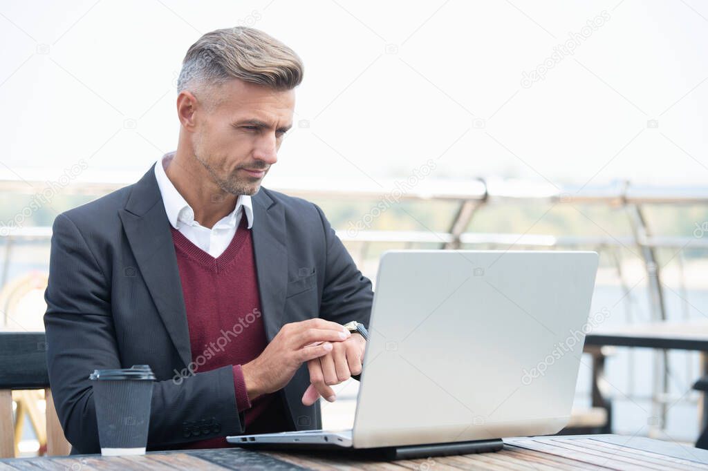 Serious professional man check time on watch working on laptop computer outside, punctuality