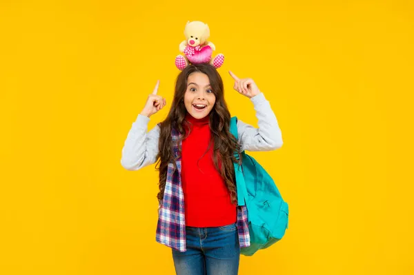 Усміхнена дівчина-підліток в повсякденному одязі тримає шкільну сумку та іграшкового ведмедя на жовтому фоні, школа — стокове фото