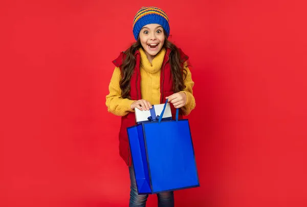 Здивована дівчина-підліток у в'язаному капелюсі тримає сумку для покупок та подарункову коробку, різдвяні покупки . — стокове фото