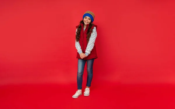 Full length of cheerful kid in knitted winter hat and down vest on red background, cold season — Stock Photo, Image