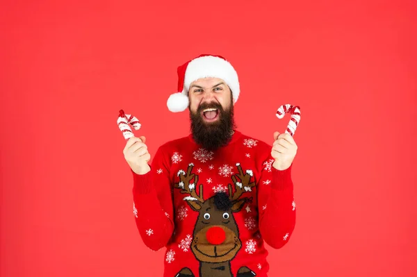 Shouting bearded man in santa claus hat and sweater hold candy stick on red background, new year — Stock Photo, Image