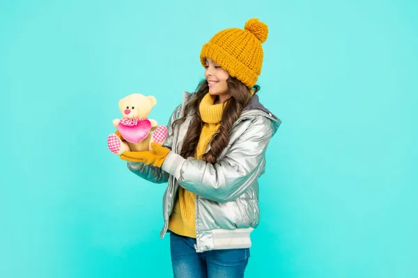 Щаслива дитина в капелюсі і пуховику з іграшкою на синьому фоні, дитинство — стокове фото
