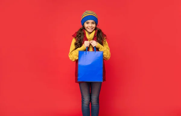 겨울 모자쓴 쾌활 한 아이가 빨간 배경으로 쇼핑 가방을 들고 크리스마스 쇼핑을 하고 있습니다. — 스톡 사진