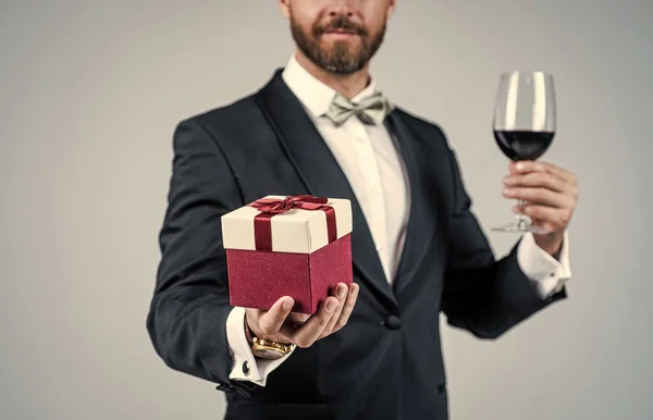 Elegant man med orakat ansikte beskuren vy i semester festlig klädsel hålla rött vin glas och presentförpackning grå bakgrund, fest — Stockfoto