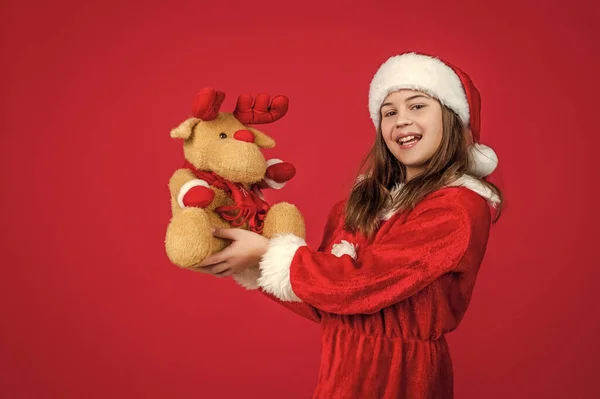Noel ziyafeti. Mutlu noeller. Neşeli genç kız Noel partisini kutluyor. Çocuk kırmızı şapka takıyor ve ren geyiği oyuncağı taşıyor. Çocuk Noel Baba kostümlü yardımcı. Parti eğlencesi. Kış tatiline hazırlanın. Mutlu yıllar. — Stok fotoğraf