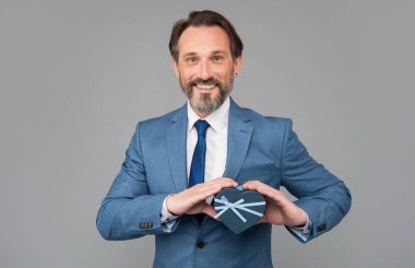 Takım elbiseli mutlu adam, gri arka planda kalp hediye kutusunu tutuyor, erkekler günü.