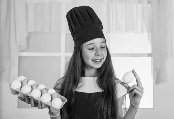 卵を用意する。幸せな子供は料理の制服を着る。帽子とエプロンのシェフの女の子。キッチンで料理を作ってる。キャリアを選択する。卵を持つ小さなヘルパー。料理も料理も。幸せな子供時代 — ストック写真