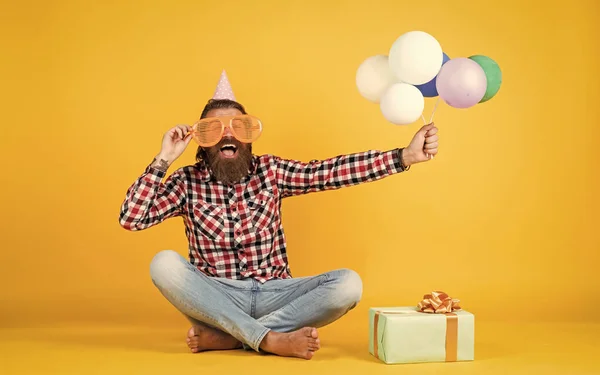 Glad födelsedagskille ser glad ut. Den skäggiga mannen känner glädjen. man med presentförpackning firar födelsedag. mogen hipster med ljusa färgglada ballonger. Galen, rolig kille. — Stockfoto