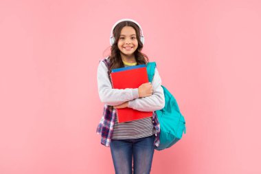 Okul çantasıyla kulaklıkla müzik dinleyen mutlu çocuk ve pembe arka plan, kitap çalışma kitabı.