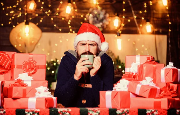 Hipster preparó regalos para la familia. Felices fiestas de invierno. Calentando. Hora del té. Hombre barbudo sombrero de Santa Claus bebiendo bebida caliente. Un año nuevo generoso. Muchos regalos. Regalos envueltos con cintas y arcos — Foto de Stock