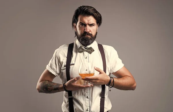 Ρετρό άντρας με παπιγιόν χαλαρώνει με αλκοόλ. αυτοπεποίθηση κομψό μπάρμαν στην κλασική φορούν πόσιμο κονιάκ. αρρενωπότητα και χάρισμα. Ουίσκι και ουίσκι. παραδοσιακός γενειοφόρος μπάρμαν — Φωτογραφία Αρχείου