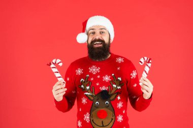 Noel Baba şapkalı ve kazaklı mutlu sakallı adam kırmızı arka planda şeker çubuğu tutuyor.