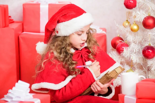 Kind santa kostuum genieten van kerstavond. Kind schrijft brief aan de kerstman. Lieve kerstman. Meisje houdt pen en papier in de buurt van kerstboom en geschenkdoos. Geloof in wonderen. Stuur een brief naar de kerstman. Wenslijst — Stockfoto