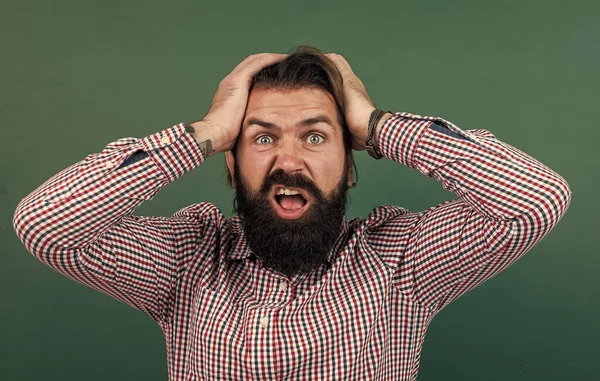 Brutal bearded man wear casual checkered shirt has headache. express human emotions. caucasian hipster on green background. shocked mature teacher on background of blackboard — Stock Photo, Image