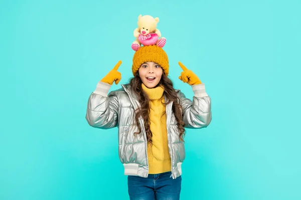 Užaslé dítě v klobouku a puffer bunda ukazováček na hračky na modrém pozadí, hračkářství — Stock fotografie