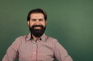 Tahtanın arkasındaki neşeli olgun öğretmen. Vahşi sakallı adam sıradan bir tişört giyiyor ve gülümsüyor. Pozitif insan duygularını ifade et. Yeşil arka planda beyaz bir hipster. Boşluğu kopyala