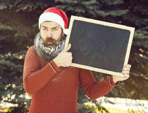Noel Baba şapkalı yakışıklı, sakallı, sakallı ve bıyıklı, beyaz donla kaplı, kışın açık havada siyah tahta tutan, doğal arka planda kaplıcayı taklit eden üzgün bir adam. — Stok fotoğraf