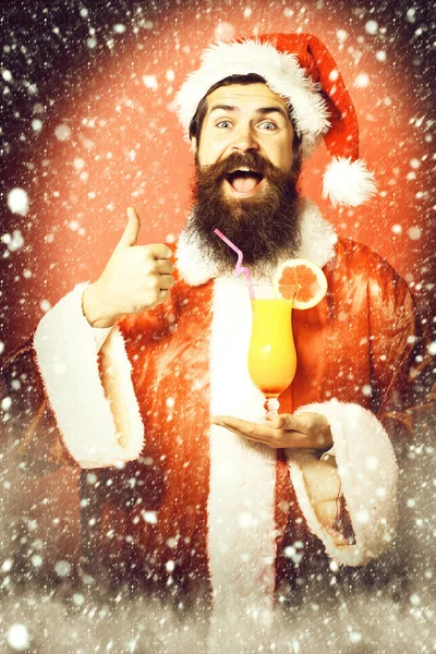 Гарний бородатий Санта-Клаус чоловік з довгою бородою на усміхненому обличчі, що тримає келих безалкогольного коктейлю в різдвяному або різдвяному светрі та новорічному капелюсі, показуючи прохолодно на червоній студії — стокове фото