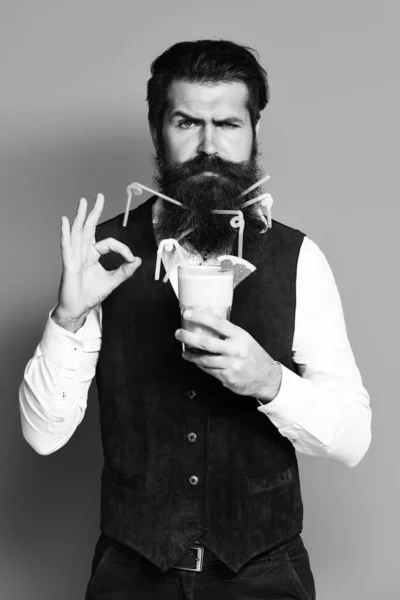 Handsome bearded man with colorful tubes in long beard and mustache has stylish hair on serious face holding glass of alcoholic cocktail in vintage suede leather waistcoat on blue studio backgrou — Fotografia de Stock