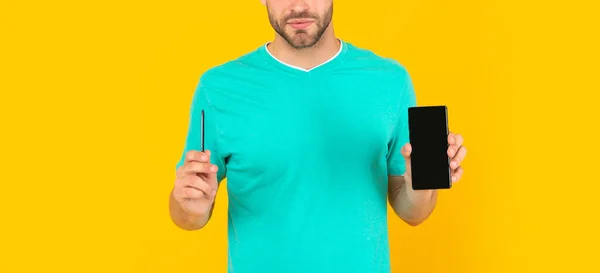 Gekappter junger Mann präsentiert Telefon und Stift auf gelbem Hintergrund, Präsentation — Stockfoto