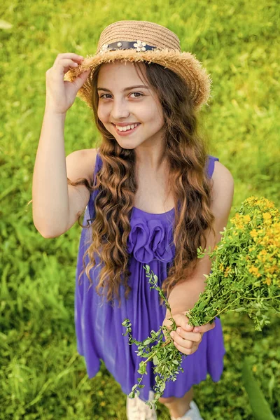 Přirozená krása. Šťastné dítě držet květiny přírodní krajiny. Holčička se usmívá se slunečním kloboukem na dlouhých vlnitých vlasech. Letní móda a doplňky. Salón krásy a lázní. Péče o pleť a vlasy — Stock fotografie