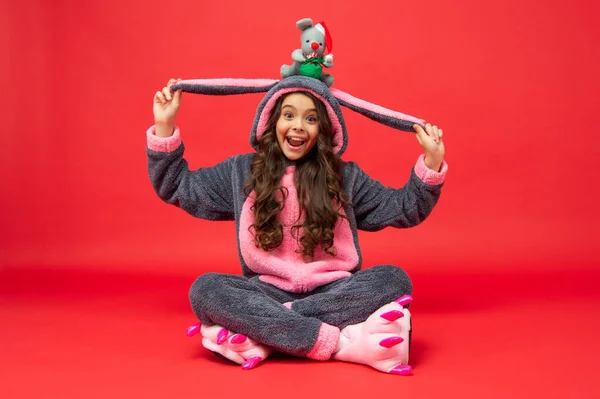 Verbaasd tiener meisje in zachte nacht pak zitten met nieuwjaar speelgoed op rode achtergrond, xmas — Stockfoto