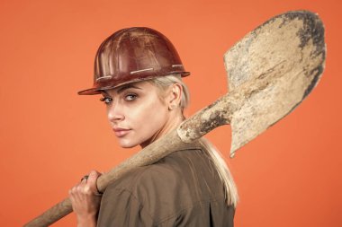 Koruyucu miğfer ve bikini giyen kadın müteahhit turuncu arka planda kürek tutuyor.