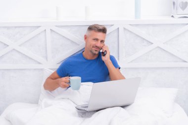 Mutlu adam cep telefonuyla konuşuyor, kahvesini yudumluyor ve sabahları laptopunu kullanıyor, iletişim kuruyor.