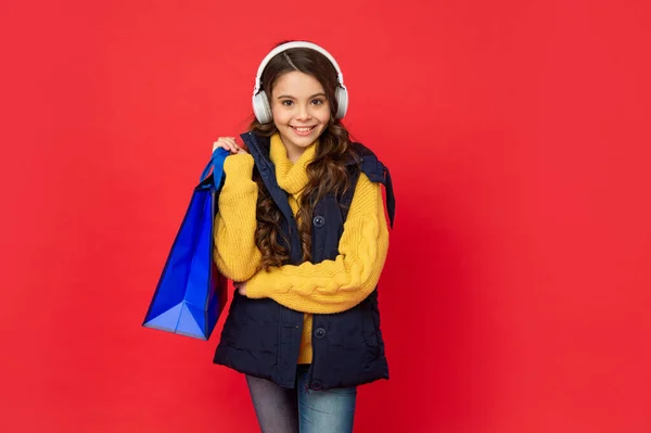 Happy kid in winter hat and earphones hold shopping bag on red background, sale — Foto Stock
