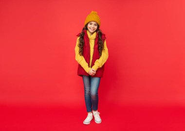 full length of happy child in knitted winter hat and down vest on red background, winter
