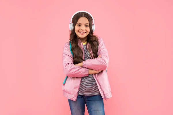 Cheerful kid listening music in earphones with school bag on pink background, music — Stockfoto