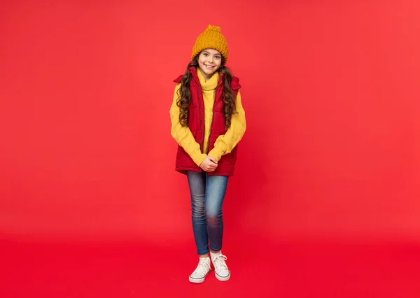 Full length of happy child in knitted winter hat and down vest on red background, winter — Stock fotografie