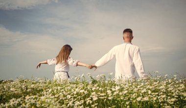 Beyaz elbiseli romantik bir çiftin, papatya çiçekleri, aşk ve özgürlük dolu bir tarlada el ele tutuşması.