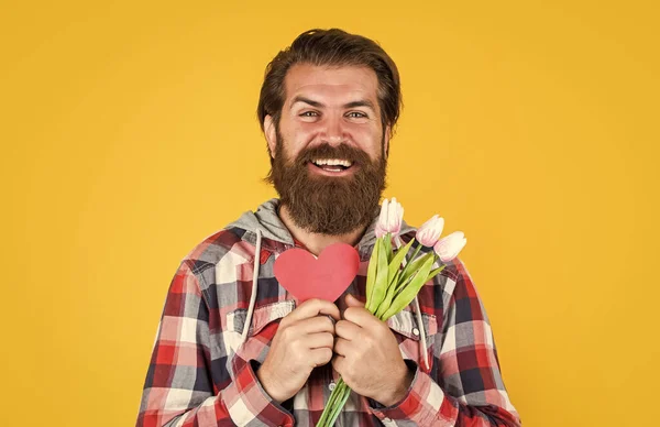 Presente de aniversário feliz com amor. Buquê de primavera para ela. Pronto para um encontro romântico. presente para mulheres e mães dia. homem barbudo feliz segurar flores de tulipa e coração decorativo. Dia dos namorados festa — Fotografia de Stock