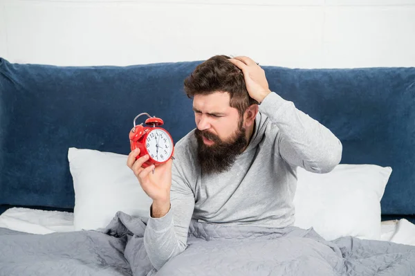 Perplexed guy scratching head holding alarm clock being in bed in morning, oversleep — Fotografia de Stock