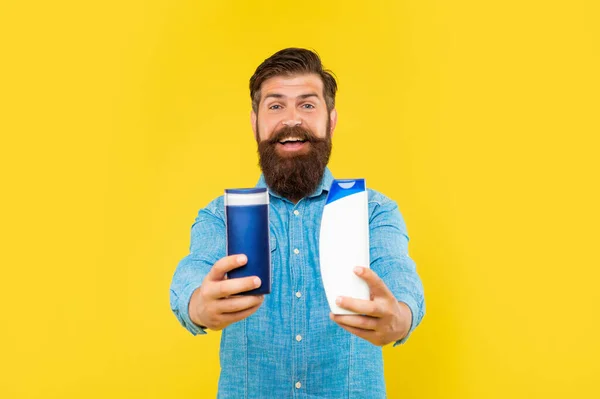 Happy brutal bearded man offer shampoo bottle on yellow background, haircare — стоковое фото