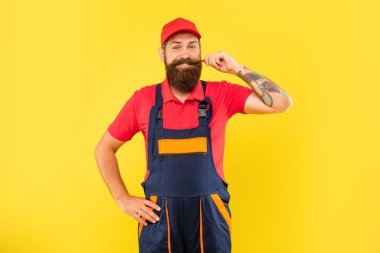 Sarı arka planda iş kıyafetleri içinde neşeli sakallı teslimatçı.