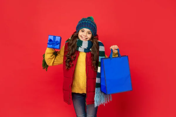 Kış şapkalı neşeli çocuk alışveriş çantası ve kırmızı arka planda hediye kutusu, boks günü. — Stok fotoğraf