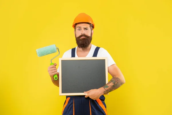 Verwirrter bärtiger Mann mit Helm und Arbeitskleidung hält Farbroller und Tafel in der Hand — Stockfoto